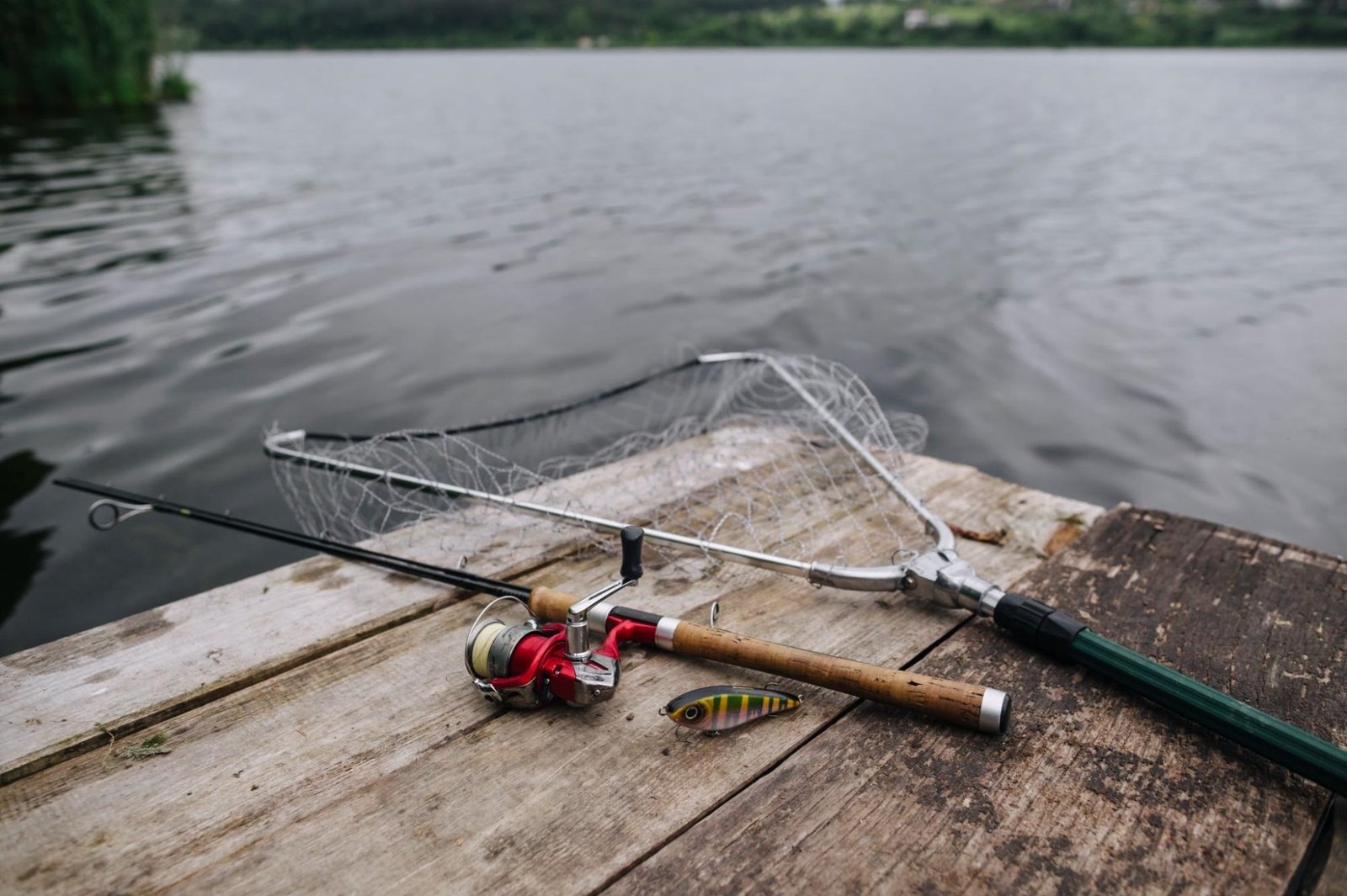 A fishing setup is arranged on a wooden dock by a calm lake. The setup includes a fishing rod with a red reel, a fishing net, and a colorful fishing lure. The background showcases lush greenery, emphasizing the peaceful environment of recreational fishing.