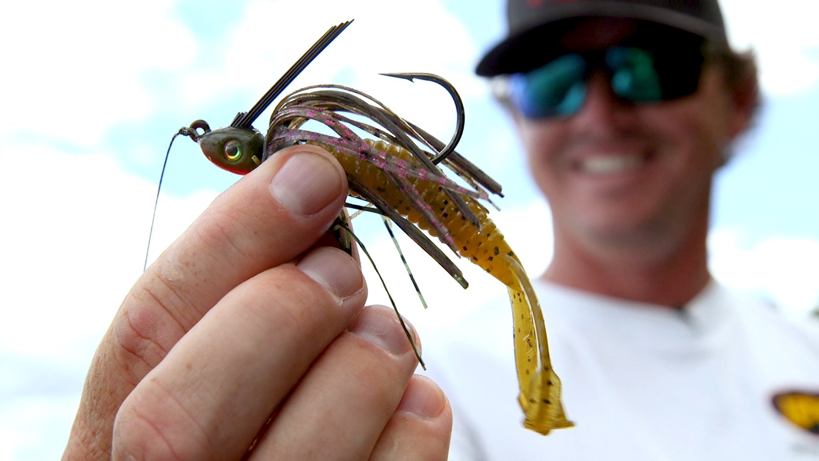 A hand holds a fishing lure designed to mimic the appearance of a small fish. The lure features a fish-shaped head with an attached hook and a body composed of multiple strands of material in yellow, brown, and pink hues. The strands give it a flowing appearance, enhancing movement in the water to attract fish.