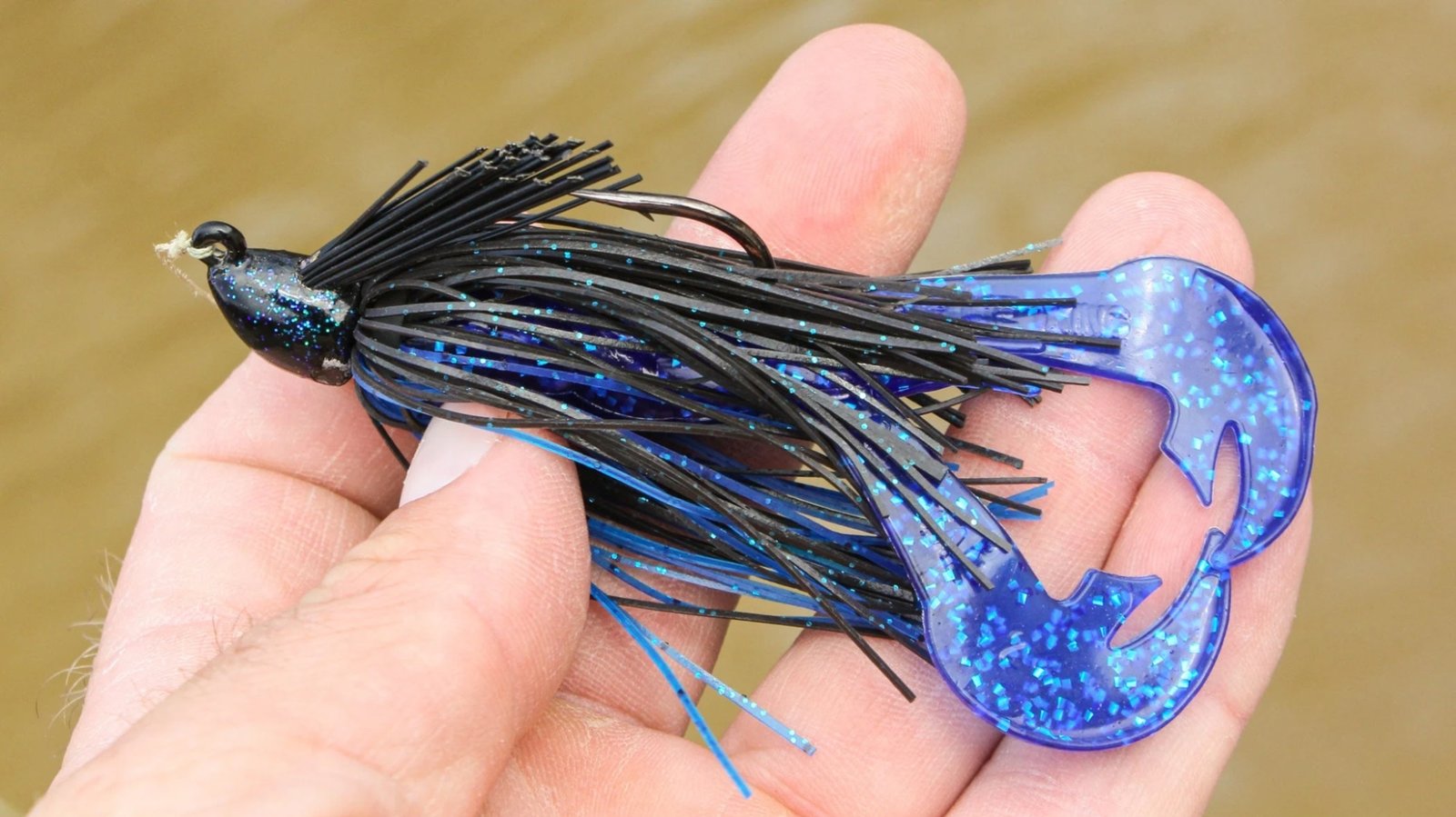A fishing lure with a black and blue color scheme featuring glittery accents is held in a person's hand. The lure has a black head with blue glitter, black and blue strands, and a blue plastic tail embedded with glitter. The tail has a unique curved design, enhancing movement in the water to attract fish.