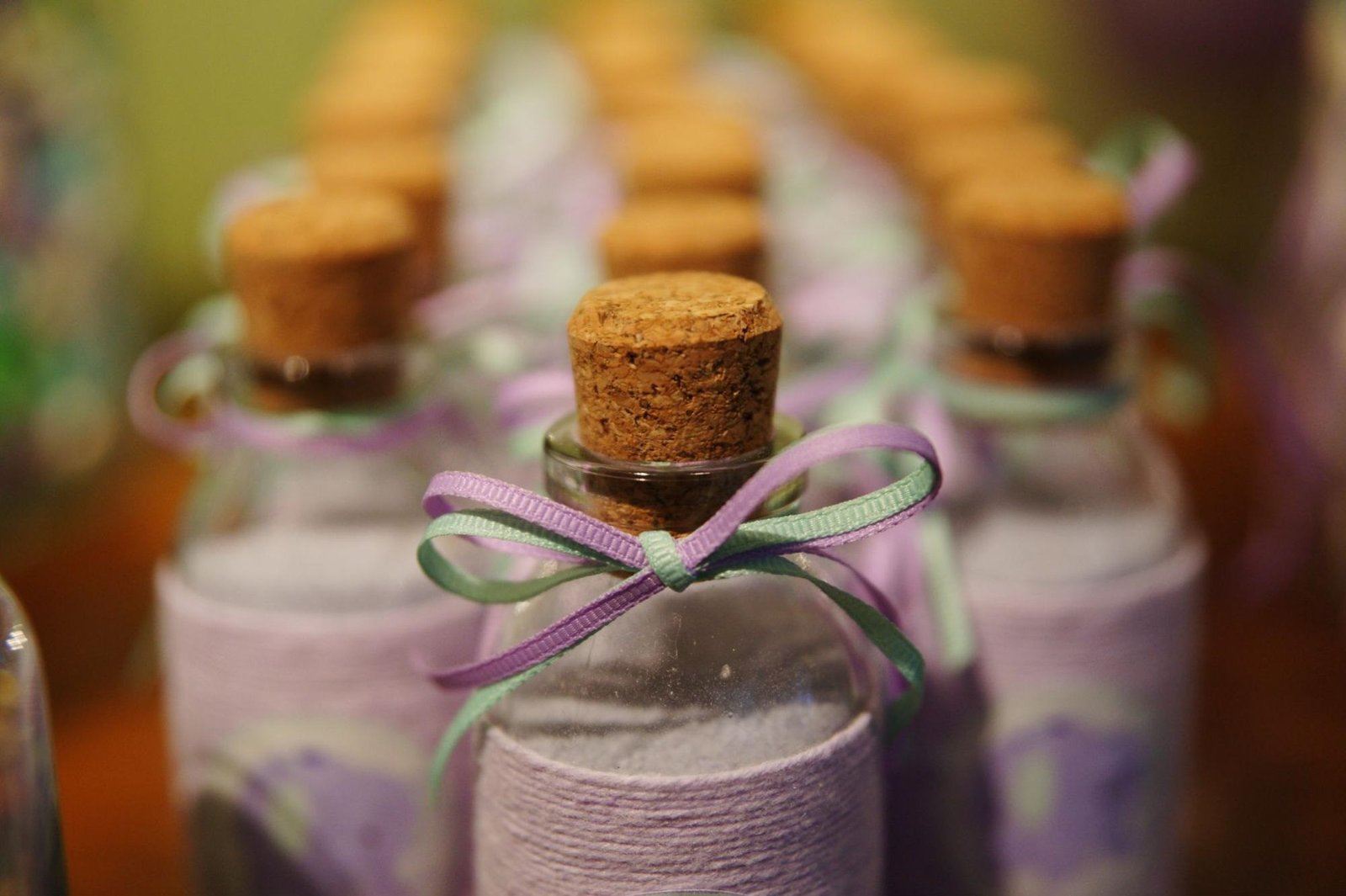 Close-up of decorative glass bottles sealed with cork stoppers, wrapped in lavender-colored fabric and tied with pastel purple and green ribbons. The bottles are arranged in neat rows, likely for a special event or as handcrafted gifts, giving a soft, elegant, and artisanal feel.